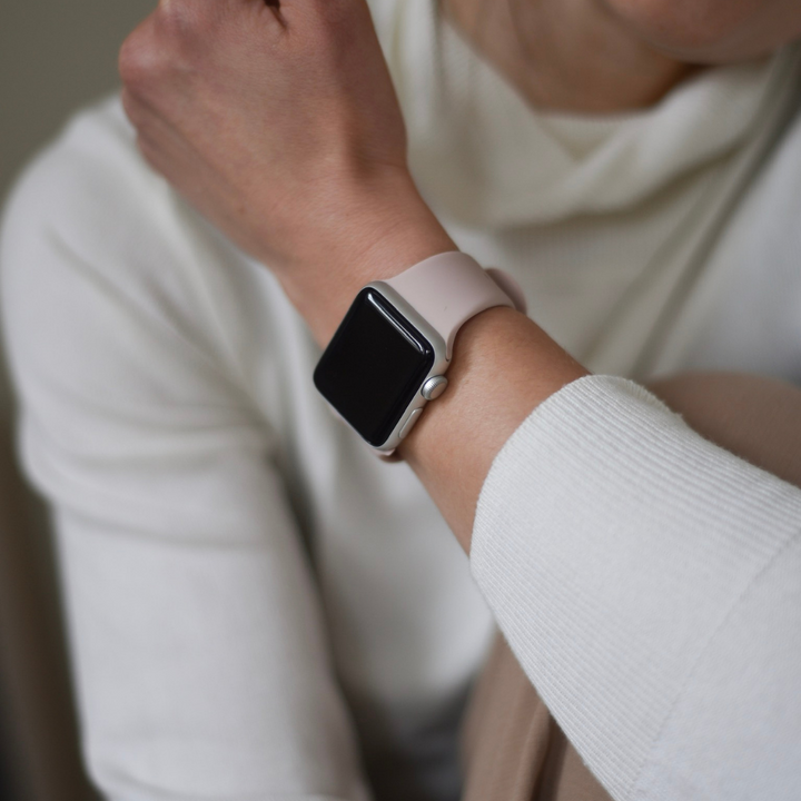 A person in a white long-sleeved shirt, with a dusty pink NAKD Apple Watch Strap by Coconut Lane on their wrist, is adjusting the watch. The smartwatch, featuring a blank dark screen and durable yet chic silicone strap, directs attention to their arm as they are slightly out of frame.
