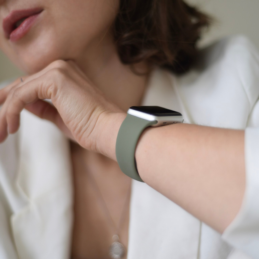 A close-up of a person wearing a white blazer and a smartwatch adorned with the NAKD Apple Watch Strap in Vintage Khaki by Coconut Lane. The person's head is partially visible, with one hand resting on their chin, drawing attention to the stylish strap on their wrist.