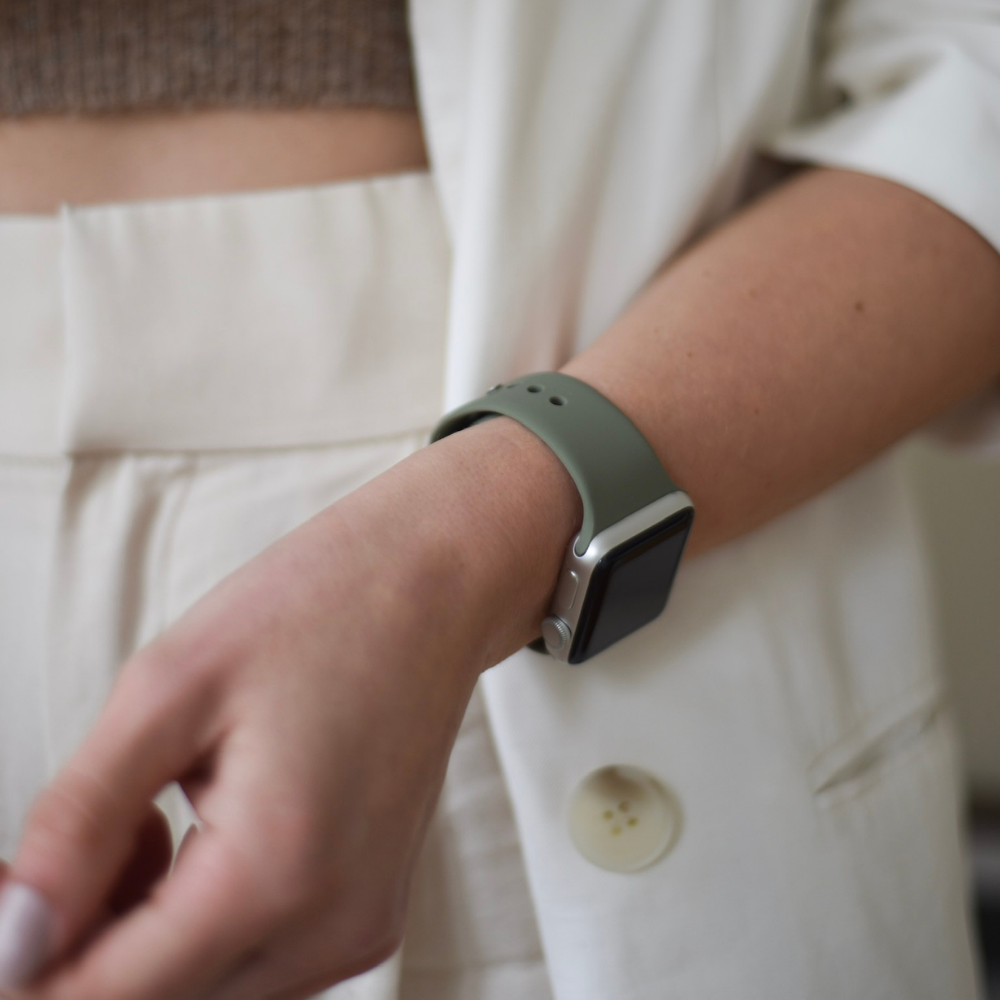 A person wearing a light green smartwatch with a NAKD Apple Watch Strap - Vintage Khaki by Coconut Lane on their wrist is seen from the torso down, dressed in a white blazer and trousers. The person is in a casual standing pose, facing forward.