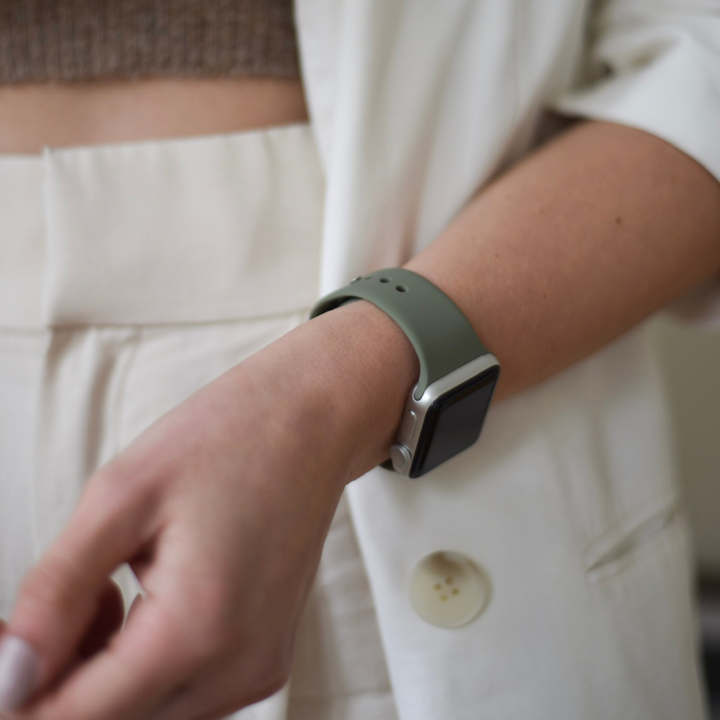A person wearing a light green smartwatch with a NAKD Apple Watch Strap - Vintage Khaki by Coconut Lane on their wrist is seen from the torso down, dressed in a white blazer and trousers. The person is in a casual standing pose, facing forward.