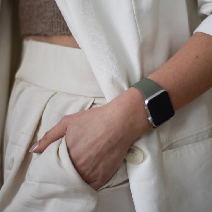 A person dressed in a white blazer and trousers is seen with one hand casually placed in their pocket. Adorning their wrist is a smartwatch with a dark screen, complemented by Coconut Lane's NAKD Apple Watch Strap in Vintage Khaki. The ensemble also includes a beige top worn under the blazer. The image emphasizes the lower torso and wrist area.