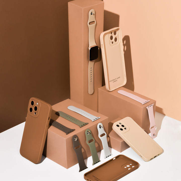 A display of various beige and brown phone cases and watch bands, including the NAKD Apple Watch Strap in Dusty Pink by Coconut Lane, arranged on beige and brown blocks. The composition includes different angles, with some items standing vertically and others lying flat. The background is a gradient of beige and brown tones.