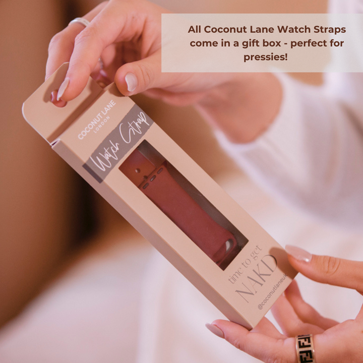 A person holding a Coconut Lane NAKD Apple Watch Strap package. The durable and chic beige box features a viewing window revealing a dusty pink silicone strap inside. Text overlay in the top right corner reads, "All Coconut Lane Watch Straps come in a gift box - perfect for pressies!