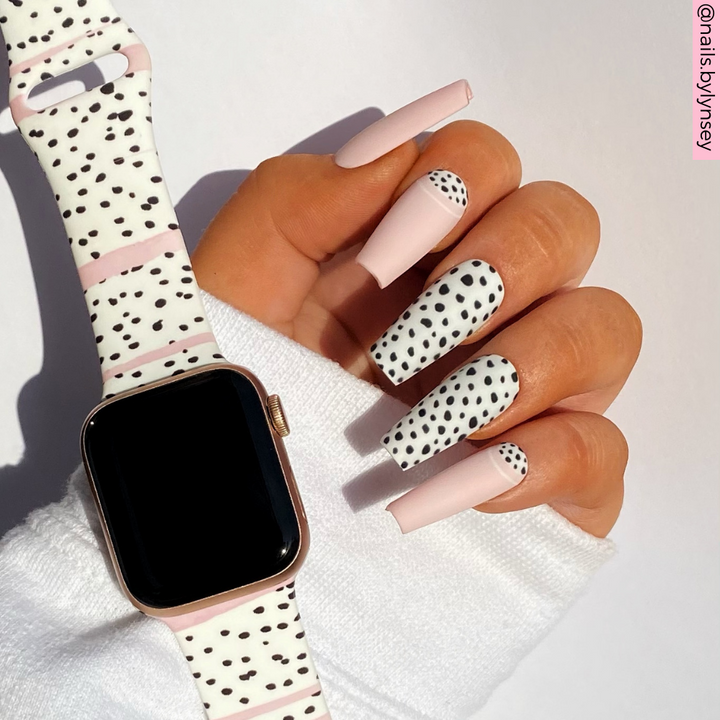A hand with long, stylish nails featuring a pink and black polka dot design is shown. The wrist flaunts an Apple Watch adorned with a Coconut Lane Pink Dalmatian Apple Watch Strap. The background is white, and the photo is bright and well-lit.