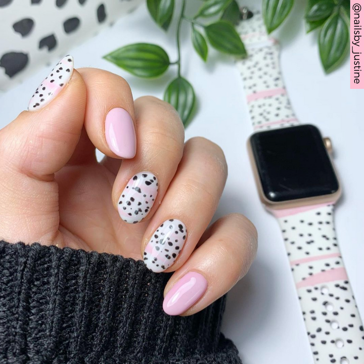 A hand with manicured nails painted in a mix of pale pink and white with black polka dots is shown. The person is wearing a black sweater. Nearby is the Coconut Lane Pink Dalmatian Apple Watch Strap, decorated with black spots to match the manicure. Green leaves are in the background. The handle "@nailsby_justine" is visible in the top right.