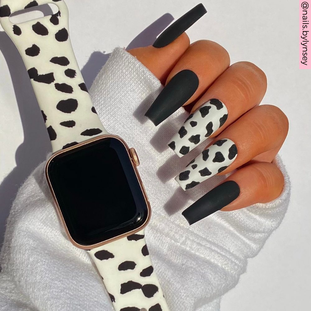 A hand adorned with long, matte black and cow-print nails is depicted in the image. The individual is dressed in a white long-sleeve top and sporting an Apple Watch fitted with Coconut Lane's Monochrome Spots Apple Watch Strap. The backdrop is a plain white, and along the right edge of the image, the Instagram handle @nailsbylynsey can be seen.