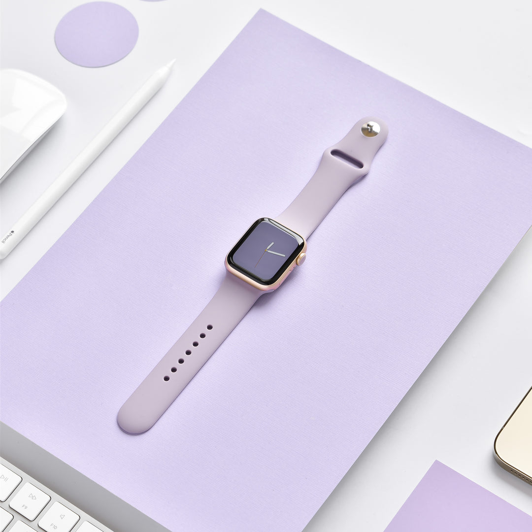 A smart watch with a Coconut Lane NAKD Apple Watch Strap in Lavender Lilac lies on a matching lavender surface, surrounded by a white stylus, a white keyboard, and circular lavender stickers. The watch display shows the time with a simple clock face. The background is minimalist and monochromatic.