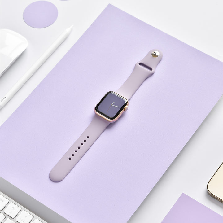 A smart watch with a Coconut Lane NAKD Apple Watch Strap in Lavender Lilac lies on a matching lavender surface, surrounded by a white stylus, a white keyboard, and circular lavender stickers. The watch display shows the time with a simple clock face. The background is minimalist and monochromatic.