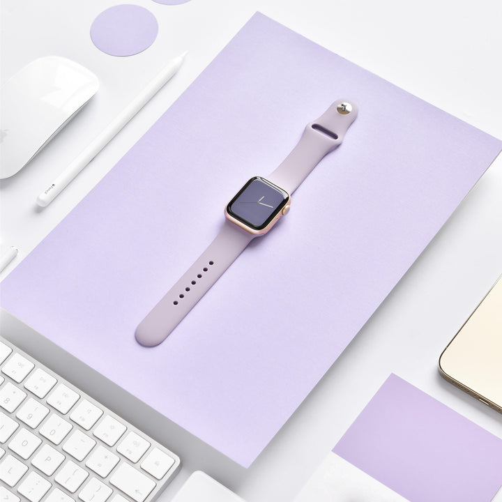 A photo showcasing a neatly arranged workspace features a Coconut Lane NAKD Apple Watch Strap in Lavender Lilac, stylishly placed on a matching lavender notebook. The setup also includes a white keyboard, white mouse, pen, and small lavender card, all set against a pristine white background.