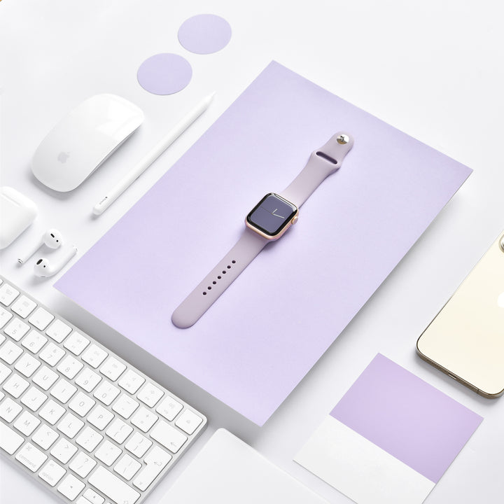 A neatly arranged flat lay of tech accessories on a white surface. Features a Coconut Lane NAKD Apple Watch Strap in Lavender Lilac centered on a lavender backdrop, along with a white keyboard, mouse, AirPods, stylus, and a gold-backed smartphone. Round lavender accents and a lavender card complete the composition.