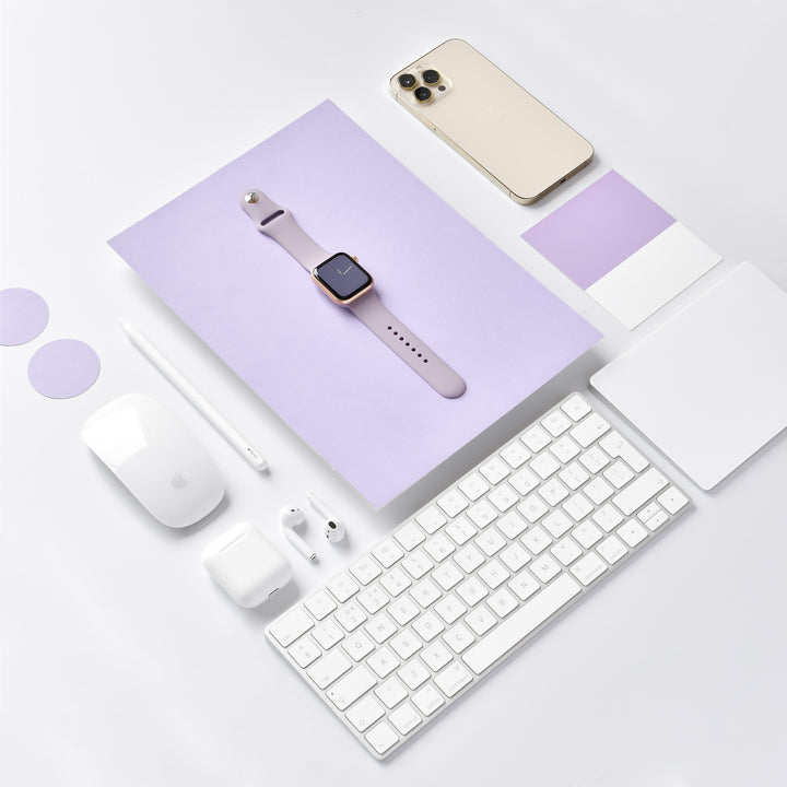 A flat lay photo showcasing the NAKD Collection by Coconut Lane, featuring the NAKD Apple Watch Strap in Lavender Lilac, an iPhone with a gold back, an Apple keyboard, a Magic Mouse, a Magic Trackpad, AirPods, a stylus, and various lavender-colored swatches and circular objects arranged neatly on a white surface.