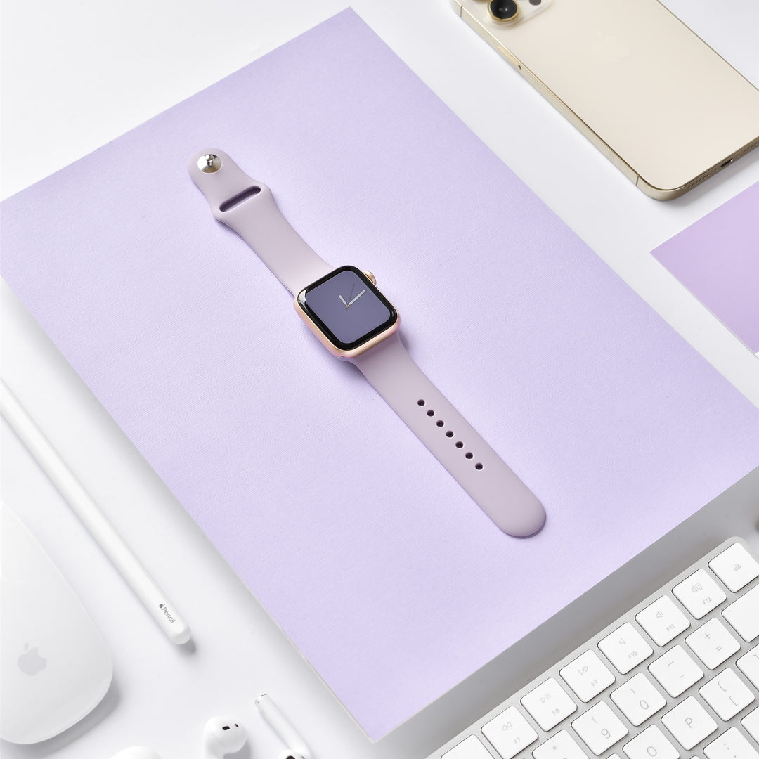 Image of a sleek smartwatch with a NAKD Apple Watch Strap - Lavender Lilac by Coconut Lane, placed on a light purple surface. Surrounding it are various tech gadgets, including a gold smartphone, a white keyboard, a white mouse, white earphones, and a white stylus.