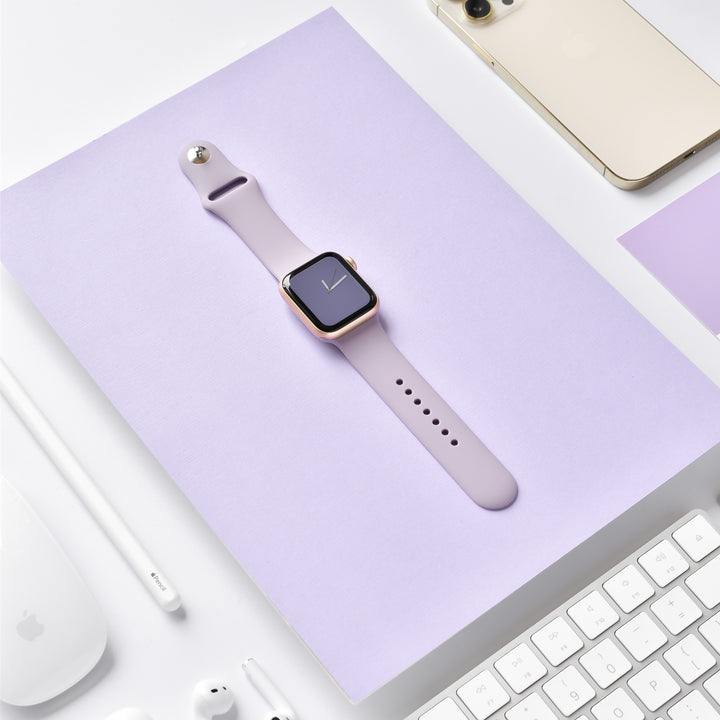 Image of a sleek smartwatch with a NAKD Apple Watch Strap - Lavender Lilac by Coconut Lane, placed on a light purple surface. Surrounding it are various tech gadgets, including a gold smartphone, a white keyboard, a white mouse, white earphones, and a white stylus.