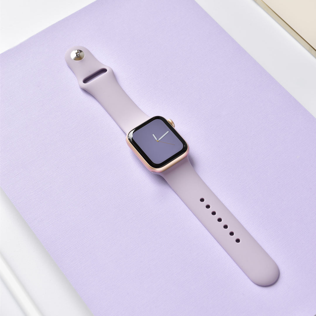 A Lavender Lilac NAKD Apple Watch Strap from Coconut Lane is displayed on a lavender-colored surface. The watch face, black with a minimalistic clock display featuring white hands, seamlessly blends into the stylish appearance of the NAKD Collection.