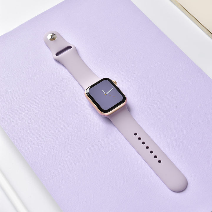 A Lavender Lilac NAKD Apple Watch Strap from Coconut Lane is displayed on a lavender-colored surface. The watch face, black with a minimalistic clock display featuring white hands, seamlessly blends into the stylish appearance of the NAKD Collection.