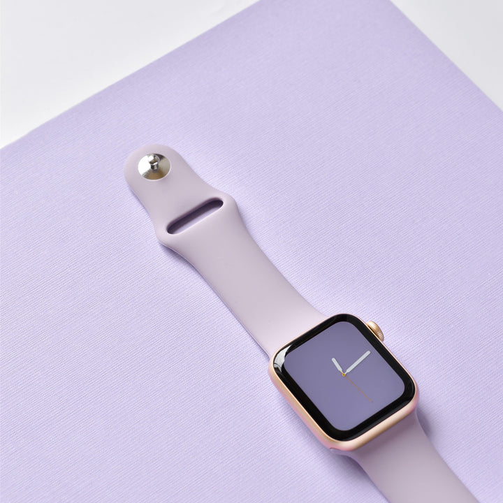 A rose gold smartwatch paired with the NAKD Apple Watch Strap - Lavender Lilac from Coconut Lane is displayed on a matching lavender surface. The screen shows a simple analog clock face with yellow hour and minute hands, while the background features a subtle texture that adds depth to the minimalist composition.