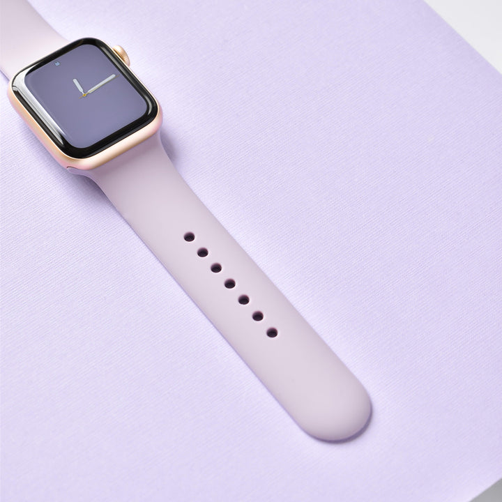 A NAKD Apple Watch Strap - Lavender Lilac from Coconut Lane, featuring a gold case and lavender band, is displayed on a pastel purple surface. The watch face shows an analog clock with a minimalist design, creating a stylish and modern aesthetic.