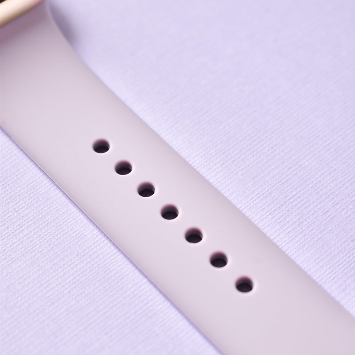 A close-up view of the Coconut Lane NAKD Apple Watch Strap - Lavender Lilac, featuring a silicone material and a gold buckle, lying on a textured, light purple surface. The strap has multiple adjustment holes for sizing.