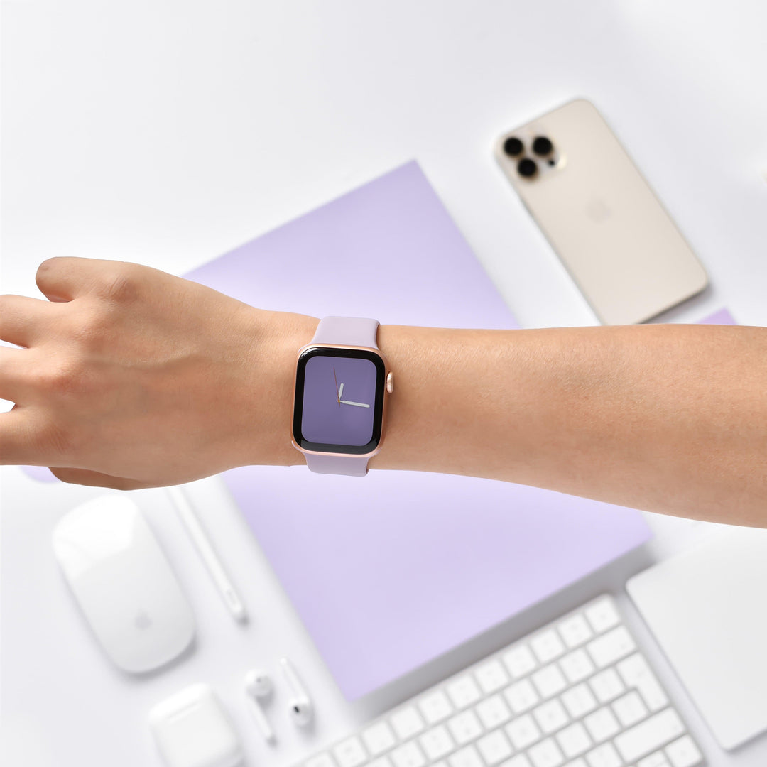 A person's left wrist showcasing a modern smartwatch with a Lavender Lilac NAKD Apple Watch Strap by Coconut Lane and a simple clock face. In the background, an organized desk setup features a gold smartphone, a keyboard, a mouse, and other tech accessories on a white and lavender surface.
