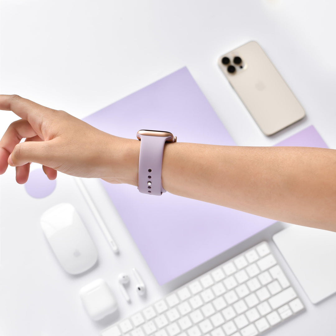 A person's wrist wearing a Lavender Lilac NAKD Apple Watch Strap from Coconut Lane is seen over a desk with tech accessories, including a smartphone, keyboard, mouse, stylus, earphones in a case, and a mouse pad, all in matching white and beige. The background features light purple paper.