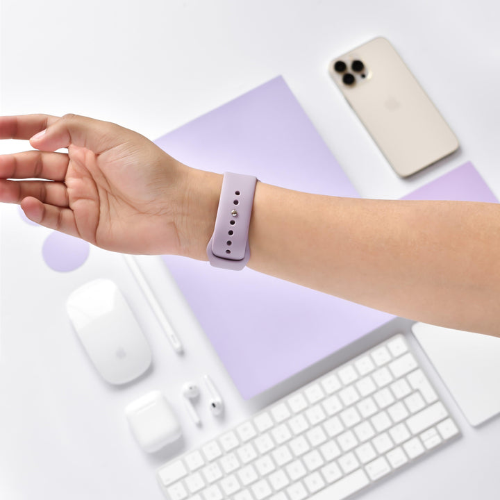 A person's arm wearing a Coconut Lane NAKD Apple Watch Strap in Lavender Lilac extends over a lavender-themed workspace. The desk features a lavender mouse mat, white mouse, keyboard, earbuds, stylus, and smartphone. The overall aesthetic is minimalistic and coordinated in pastel hues from the NAKD Collection.