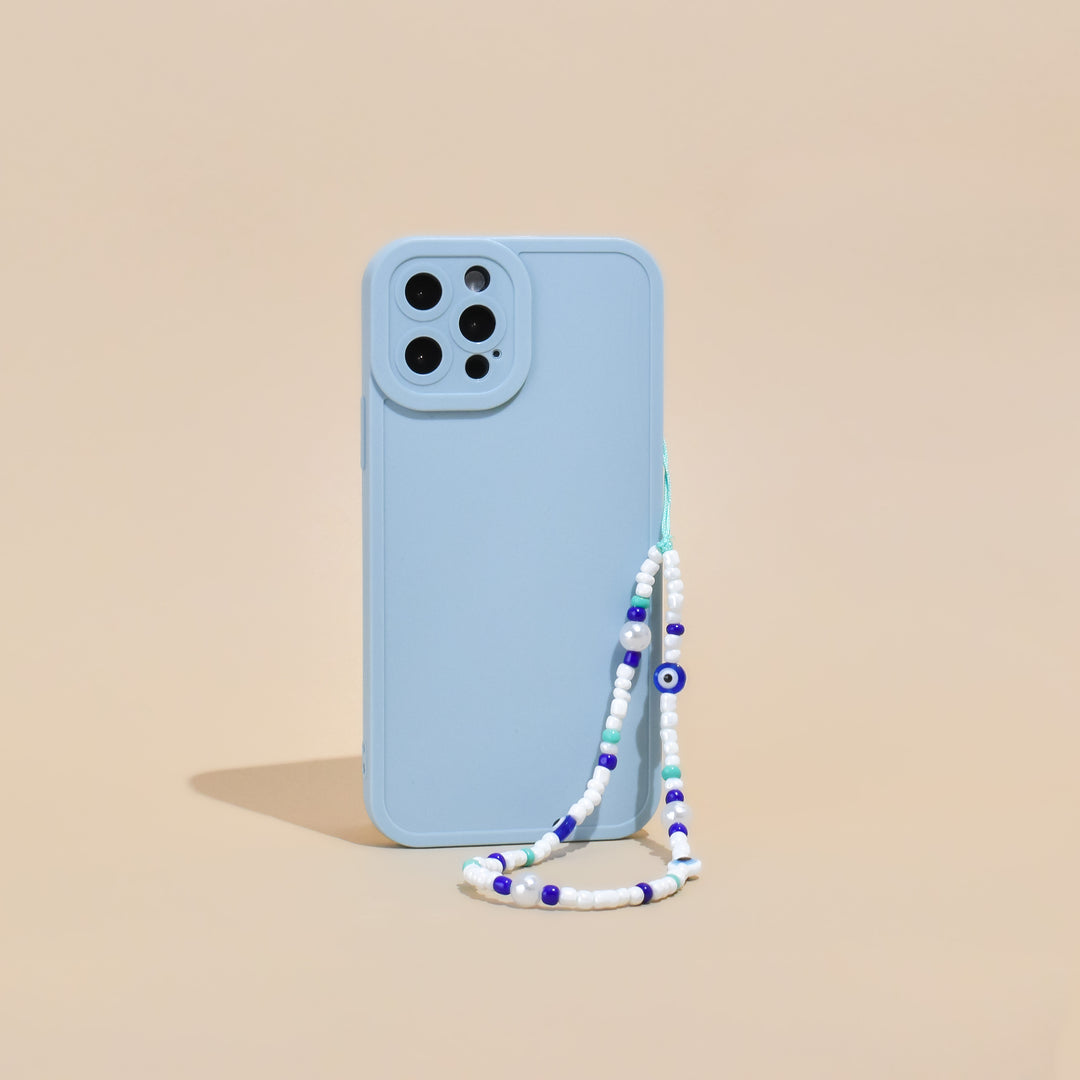 A light blue smartphone case with a Coconut Lane Beaded Phone Strap - Evil Eye is standing upright against a beige background. The beaded strap features blue, white, and purple beads, including one with an eye design. The setup boasts a minimalist aesthetic.