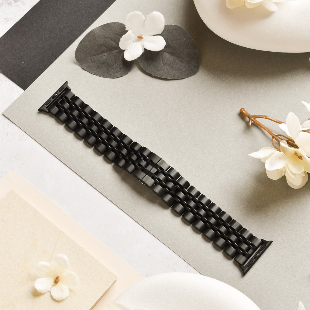 The Stainless Steel Apple Watch Strap in Black by Coconut Lane is displayed on a light gray surface, surrounded by white flowers as well as gray and beige decorative papers. The scene exudes a minimalist and elegant aesthetic, with soft lighting emphasizing the textures of the adjustable strap and surrounding elements.