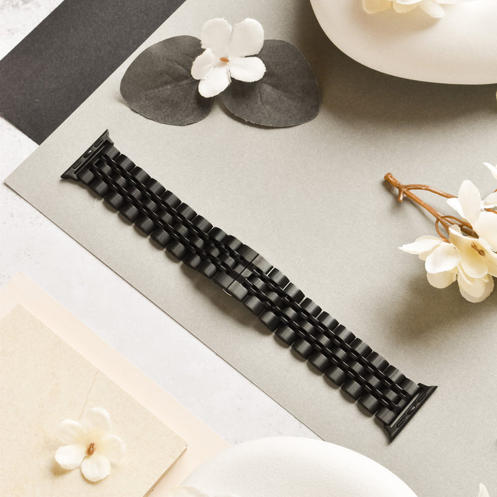 The Stainless Steel Apple Watch Strap in Black by Coconut Lane is displayed on a light gray surface, surrounded by white flowers as well as gray and beige decorative papers. The scene exudes a minimalist and elegant aesthetic, with soft lighting emphasizing the textures of the adjustable strap and surrounding elements.