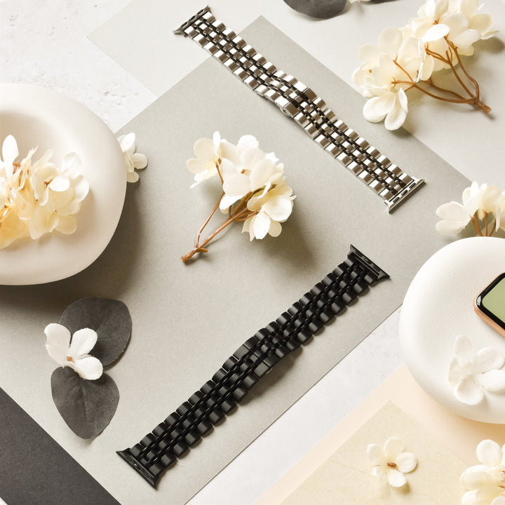 Two watch bands, one silver and one black, are displayed on a flat surface adorned with white flowers and green leaves. A phone partially visible on the right side adds a touch of modernity to the elegant floral arrangement. The Stainless Steel Apple Watch Strap - Black by Coconut Lane fits various Apple Watch models seamlessly.