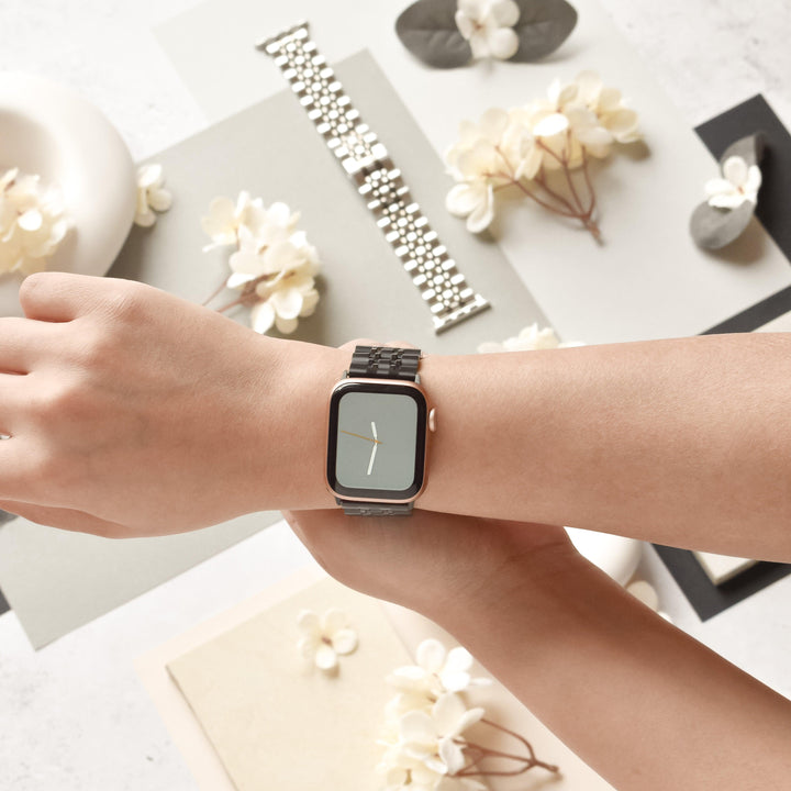 A person wearing a Coconut Lane Stainless Steel Apple Watch Strap - Black on their left wrist. White flowers, leaves, and a silver watch band are scattered on a light-colored surface in the background. The adjustable strap fits various Apple Watch models as the person holds their wrist with their right hand.