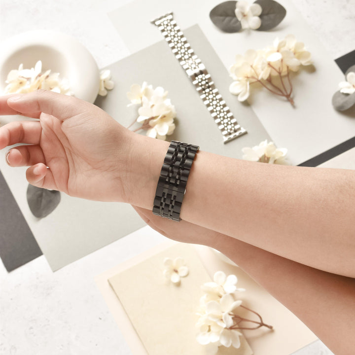 A wrist embellished with a Coconut Lane Stainless Steel Apple Watch Strap - Black stretches over a flat surface adorned with flowers and petals, contrasting grey, black, and beige paper backgrounds. A stainless steel adjustable strap rests in the background among the blossoms.