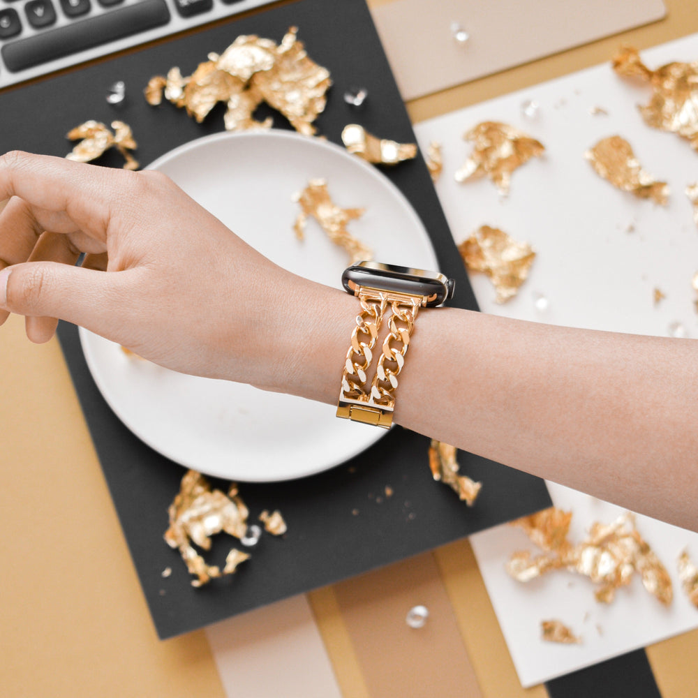 Close-up of a person wearing a stylish Dual-Chain Apple Watch Strap by Coconut Lane, featuring a thick gold chain on their wrist. The hand rests near a white plate filled with gold foil fragments. The background features a keyboard, scattered gold foil, and papers in various neutral tones.