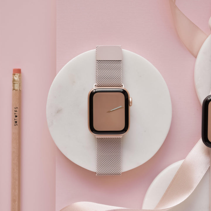 A smart watch with a dusty pink band rests on a circular marble plate, set against a pink background. To the left, there's a pencil with a pink eraser and text on the side. Part of a pink ribbon is also visible on the right. The watch features Coconut Lane’s Magnetic Chain Apple Watch Strap for added elegance and security.