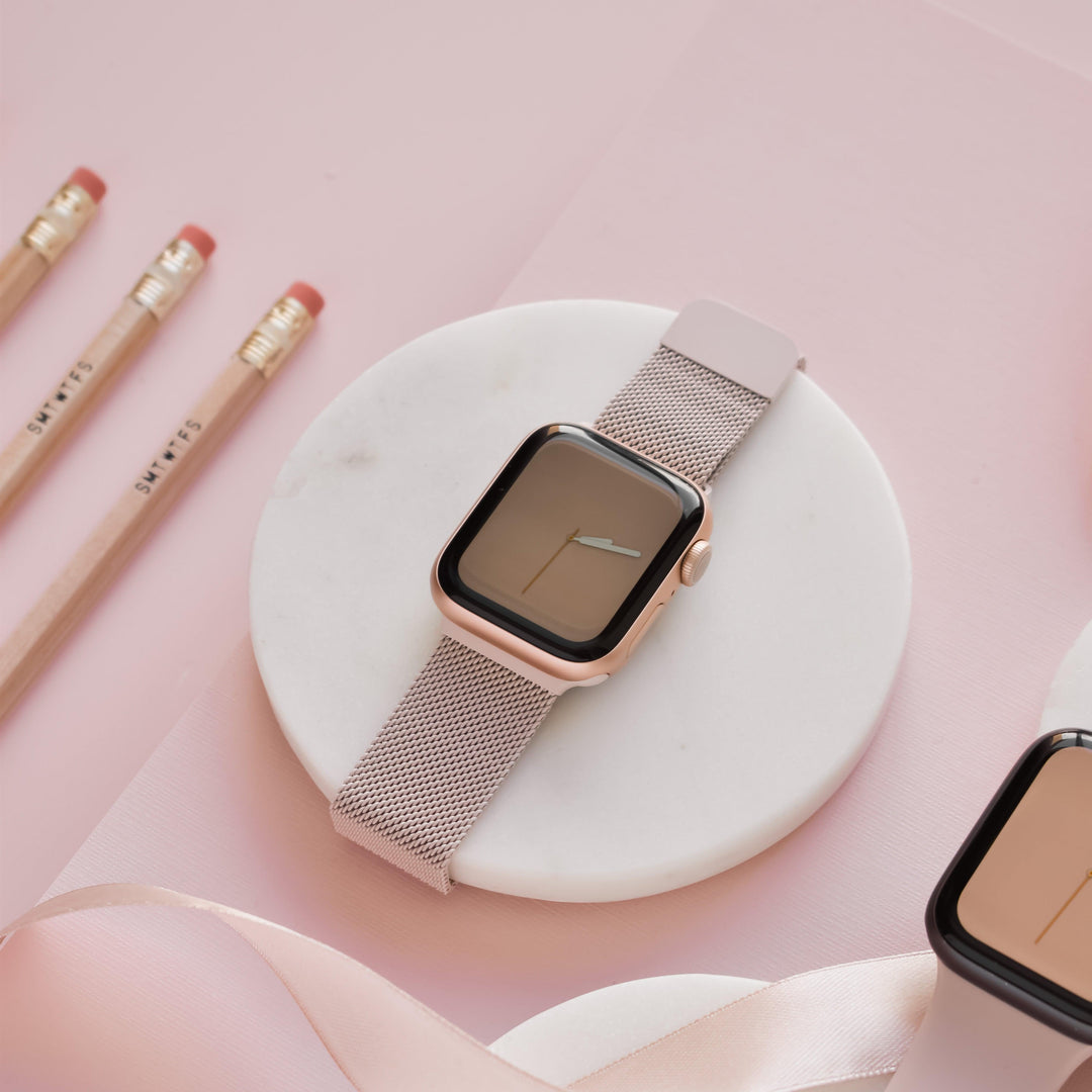A Coconut Lane Magnetic Chain Apple Watch Strap in Dusty Pink is displayed on a white circular stand against a pastel pink background. Three pencils with erasers sit to the left, partially on the surface. A piece of pink ribbon is curled in the bottom right corner.