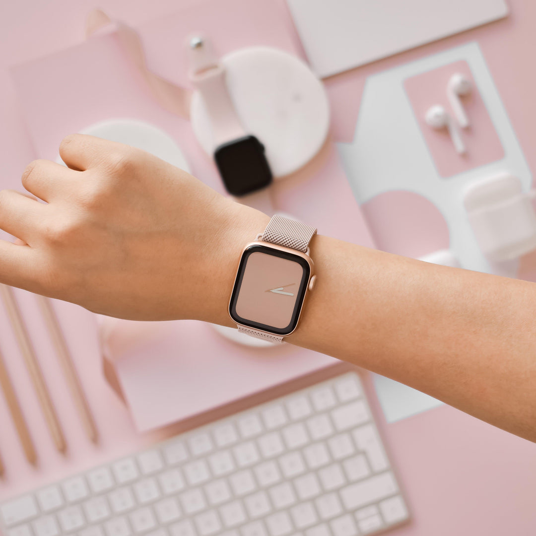 A person's wrist adorned with the Coconut Lane Magnetic Chain Apple Watch Strap in dusty pink, set against a pink and white backdrop. The scene includes a keyboard, earbuds, an additional wristwatch band, a charging dock, and several pens thoughtfully arranged for aesthetic appeal.