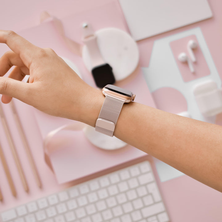 A hand wearing a Coconut Lane Magnetic Chain Apple Watch Strap in Dusty Pink is shown over a pastel pink workspace. The desk features a keyboard, stylus pens, a notepad, wireless earbuds, and a white charging dock with a second smart watch. The color scheme is predominantly pink and white.