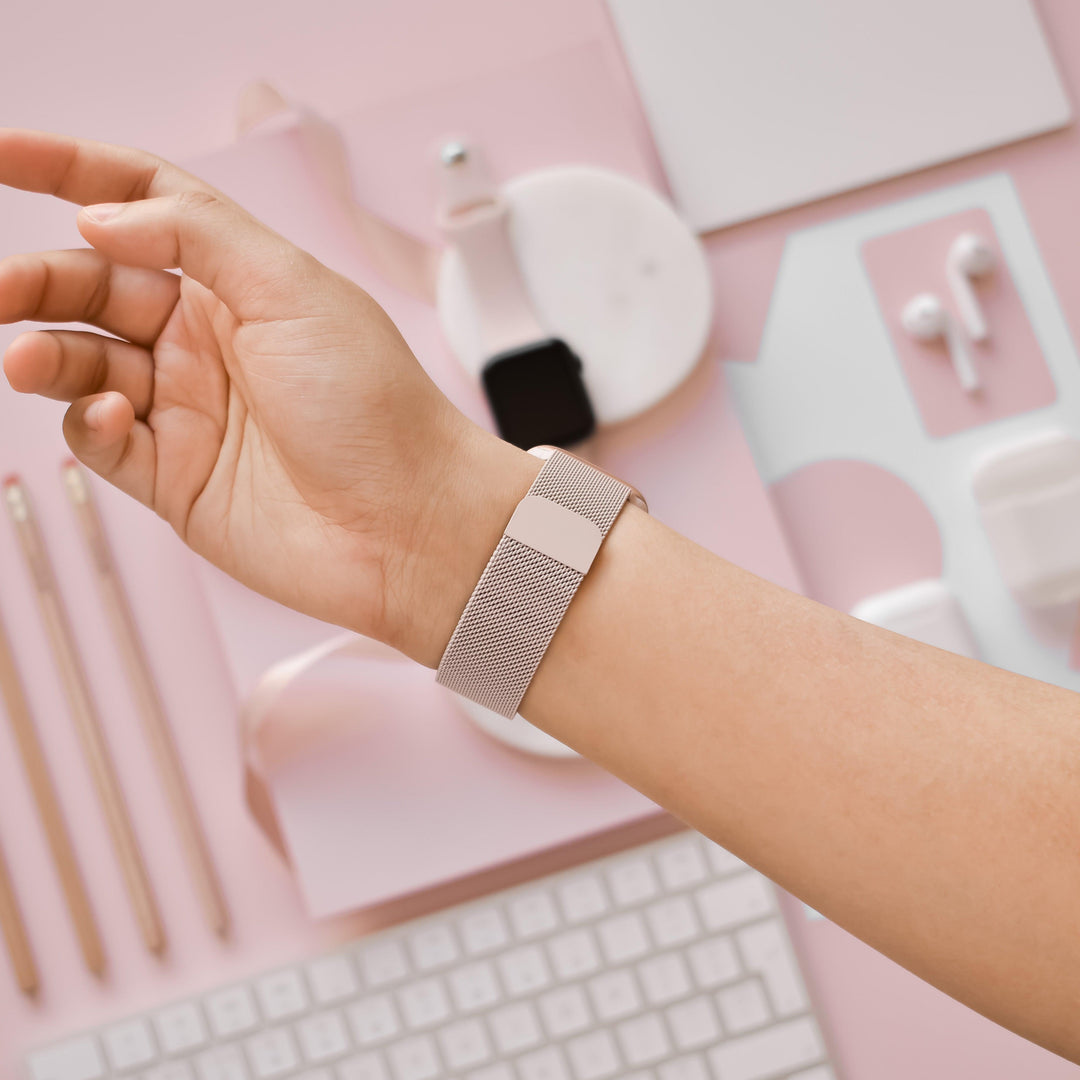 A person's arm is shown wearing a stylish Coconut Lane Magnetic Chain Apple Watch Strap in Dusty Pink on their wrist. The background includes various pink and white desk accessories such as pencils, a keyboard, a mouse, earbuds, and a small notebook, all arranged in an aesthetically pleasing manner.