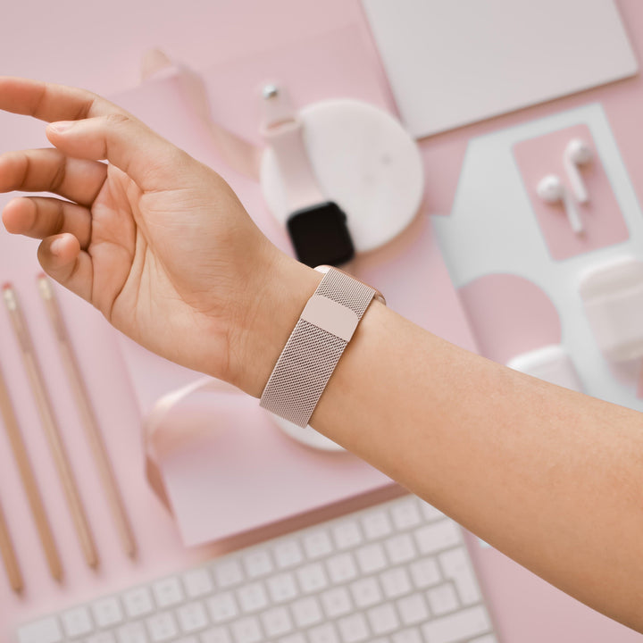 A person's arm is shown wearing a stylish Coconut Lane Magnetic Chain Apple Watch Strap in Dusty Pink on their wrist. The background includes various pink and white desk accessories such as pencils, a keyboard, a mouse, earbuds, and a small notebook, all arranged in an aesthetically pleasing manner.