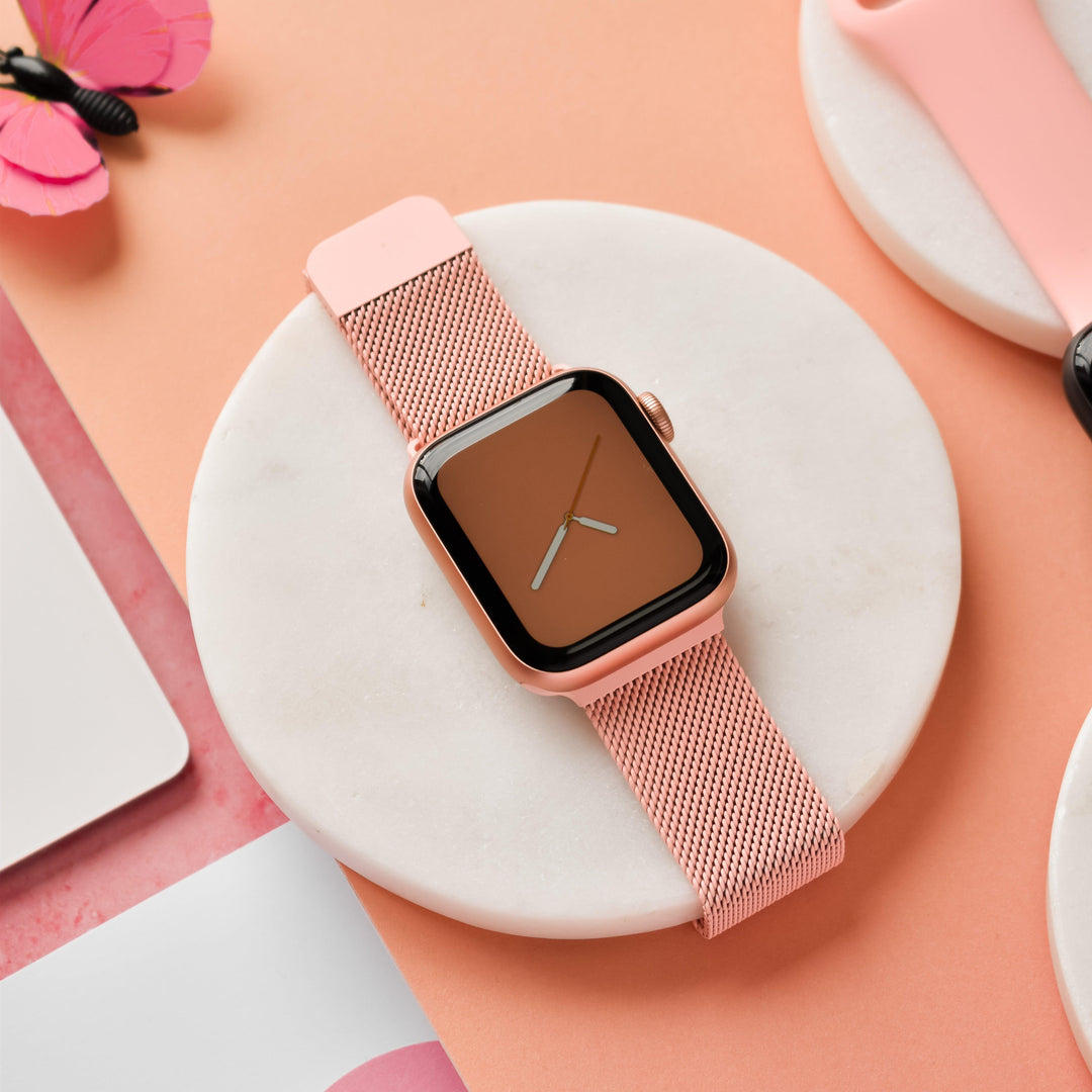 A Coral Pink Magnetic Chain Apple Watch Strap from Coconut Lane is displayed on a round white marble platform. The background features various shades of pink and coral, with a pink paper butterfly in the upper left corner. The watch screen is orange with simple, white clock hands.