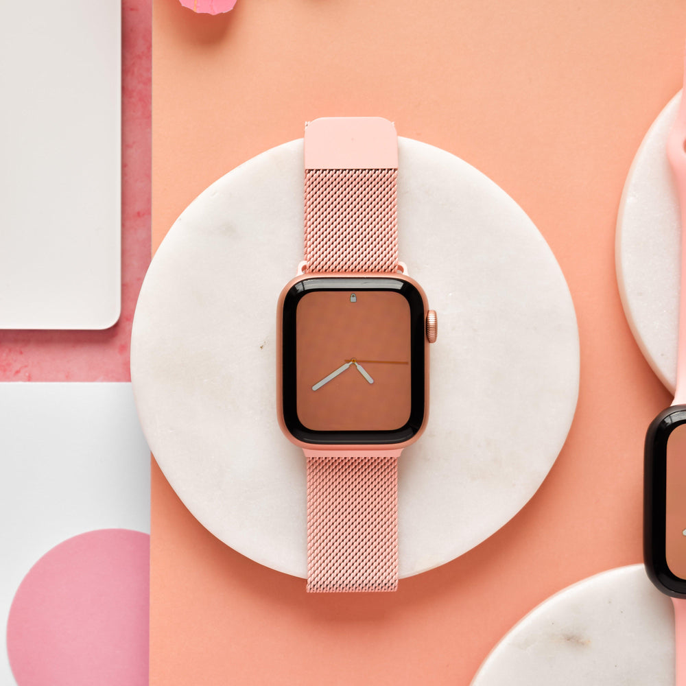 A Coral Pink smartwatch with a matching Magnetic Chain Apple Watch Strap from Coconut Lane is displayed on a round, white marble background. The watch face features a minimalistic design with a light orange background and simple white clock hands. The setting includes soft pastel colors for an elegant touch.