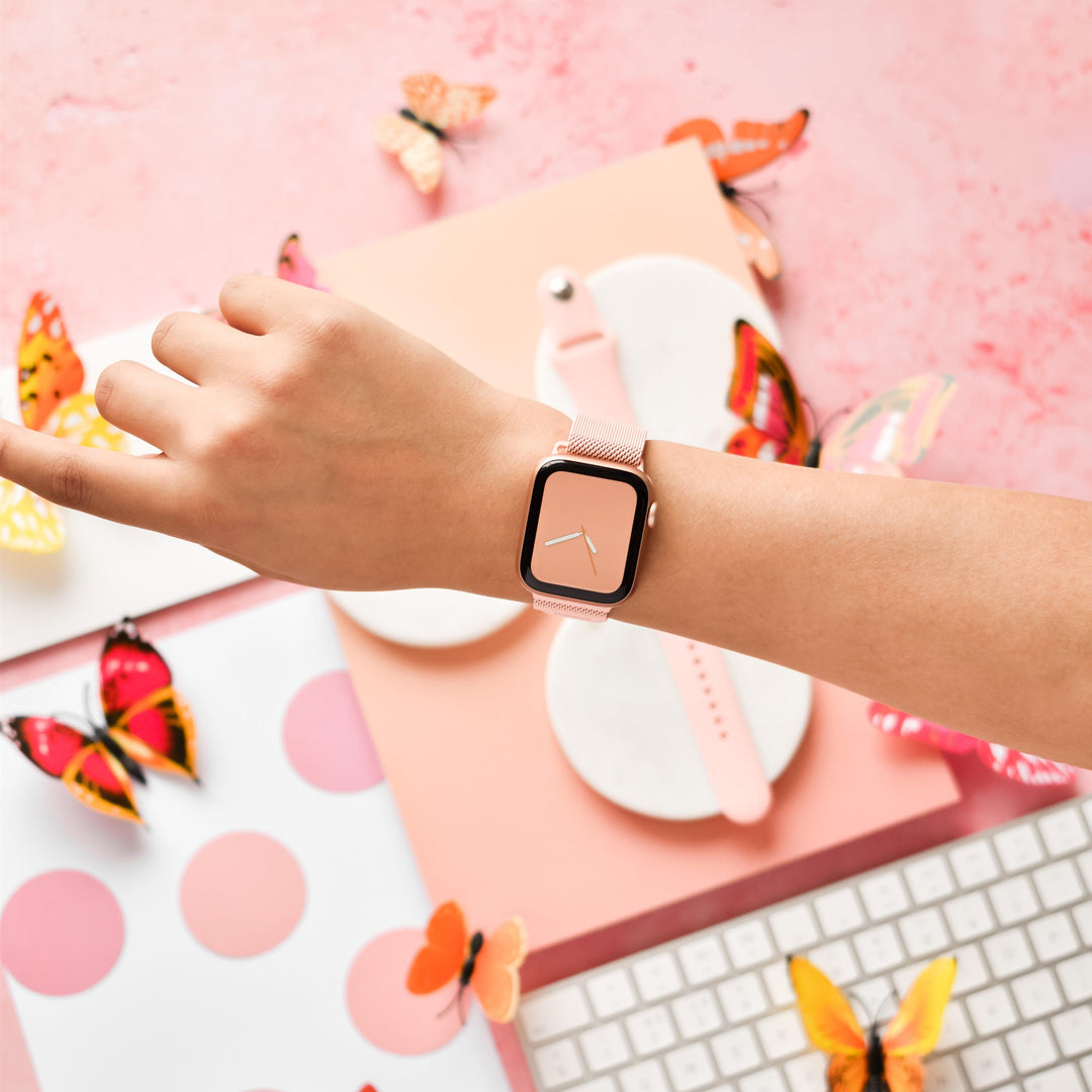 A person's wrist wearing a sleek Coconut Lane Magnetic Chain Apple Watch Strap in Coral Pink is extended into a pink-themed setting. The background includes a keyboard, a notebook with a pink polka dot cover, and scattered butterfly decorations in various colors and designs.