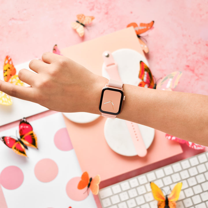 A person's wrist wearing a sleek Coconut Lane Magnetic Chain Apple Watch Strap in Coral Pink is extended into a pink-themed setting. The background includes a keyboard, a notebook with a pink polka dot cover, and scattered butterfly decorations in various colors and designs.