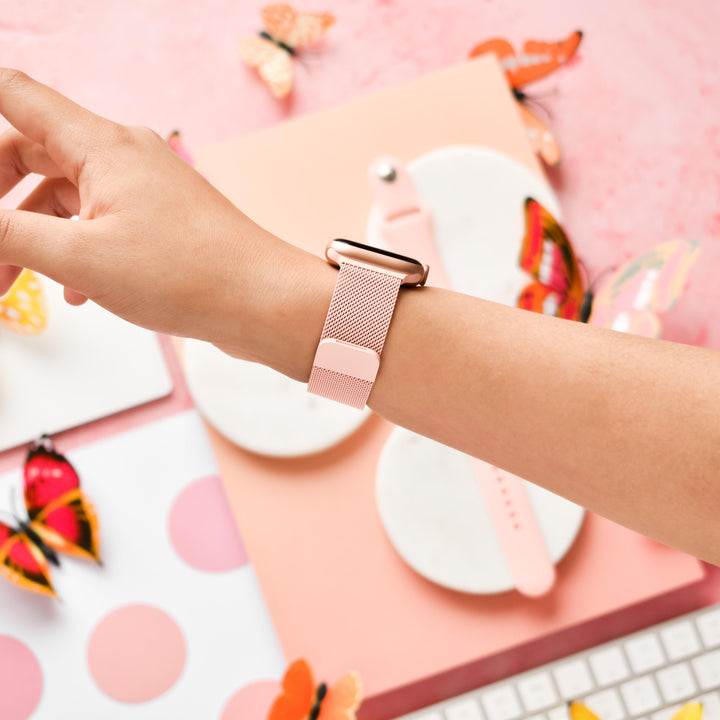 A hand wearing a Coral Pink Magnetic Chain Apple Watch Strap by Coconut Lane. The background includes colorful butterfly decorations, a pink polka dot notebook, another pink Apple Watch strap, and a portion of a keyboard. The scene is set against a soft pink surface.