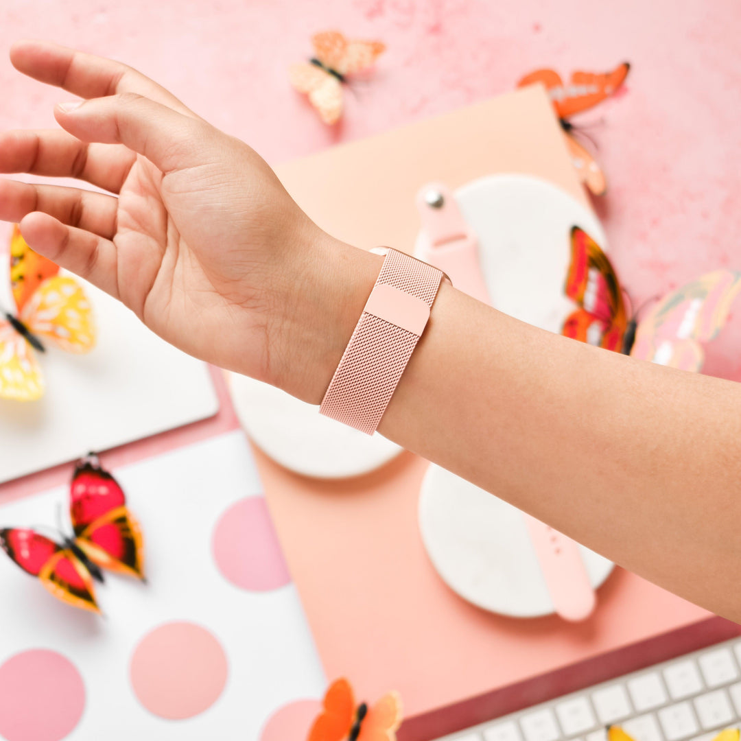 A person’s wrist is shown wearing a *Coconut Lane Magnetic Chain Apple Watch Strap in Coral Pink*. The background includes vibrant, colorful butterflies and various stationery on a pink surface, including polka dot paper, a white dish, and a keyboard. This stylish strap adds an elegant touch to the scene.