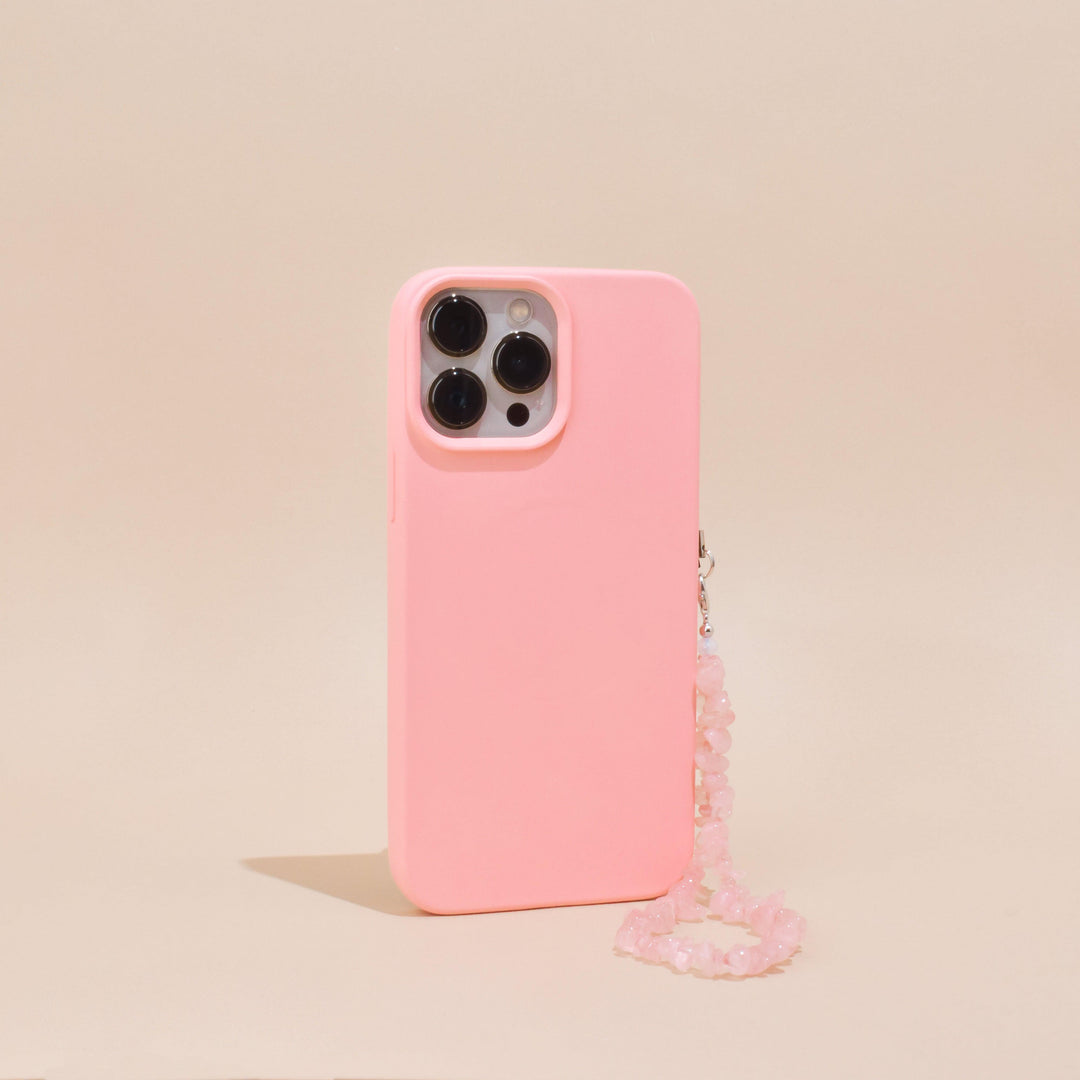A pink smartphone case featuring Coconut Lane's Crystal Phone Strap - Rose Quartz is displayed on a beige background. The case features precise cutouts for three camera lenses and a flash. The pink beaded strap, adorned with genuine rose quartz crystals, is attached to the side of the case, providing an elegant decorative touch.
