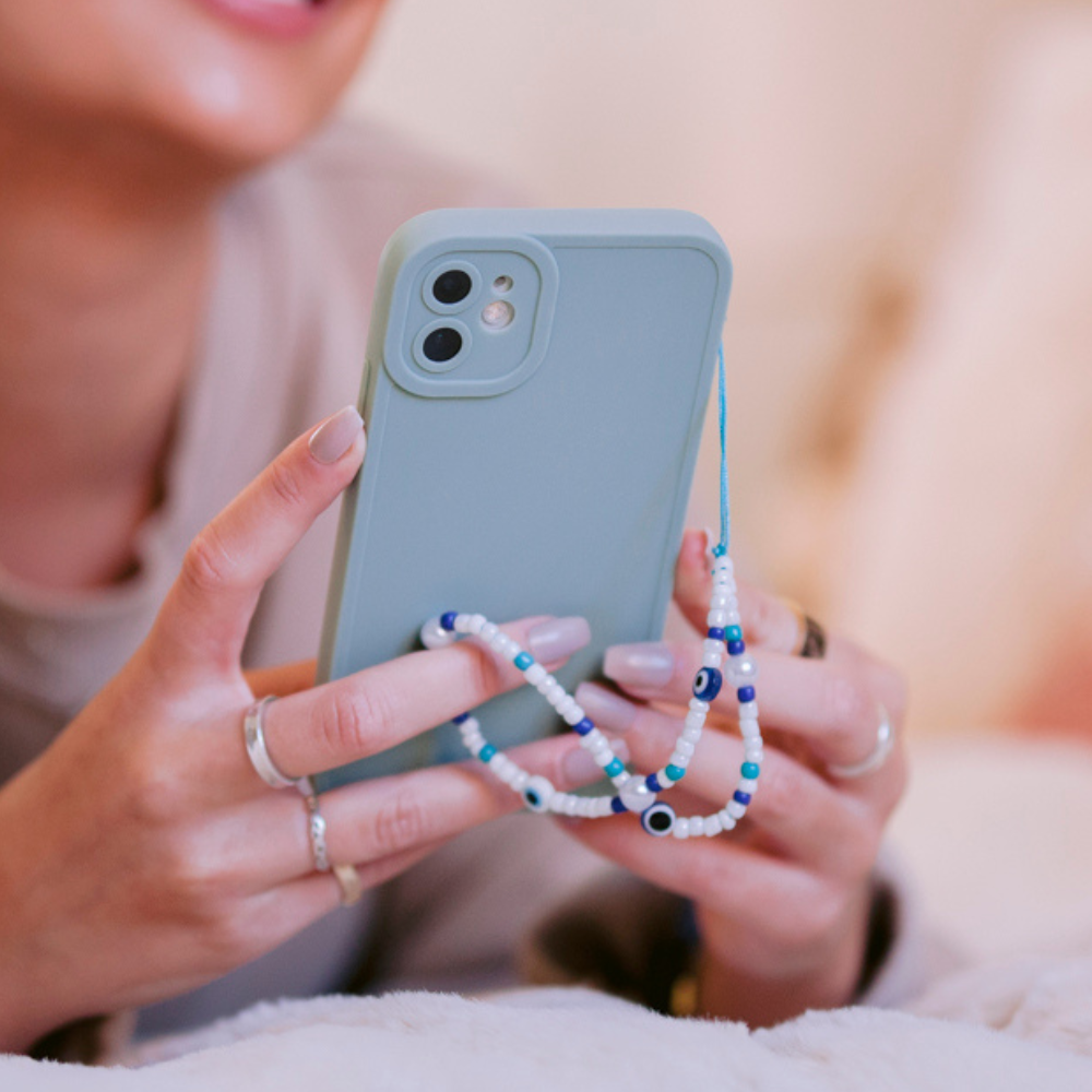 Close-up of a person holding a mint green phone adorned with the Beaded Phone Strap - Evil Eye from Coconut Lane. The person, with light skin, is wearing nail polish and multiple rings. The phone case has cutouts for two camera lenses. The background is out of focus with warm, muted tones.