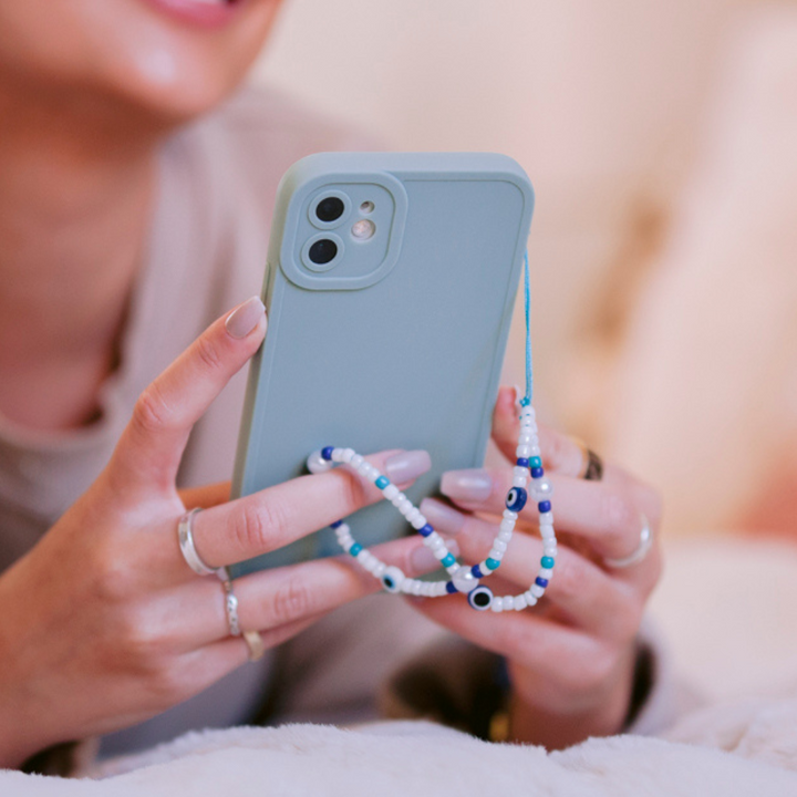 Close-up of a person holding a mint green phone adorned with the Beaded Phone Strap - Evil Eye from Coconut Lane. The person, with light skin, is wearing nail polish and multiple rings. The phone case has cutouts for two camera lenses. The background is out of focus with warm, muted tones.