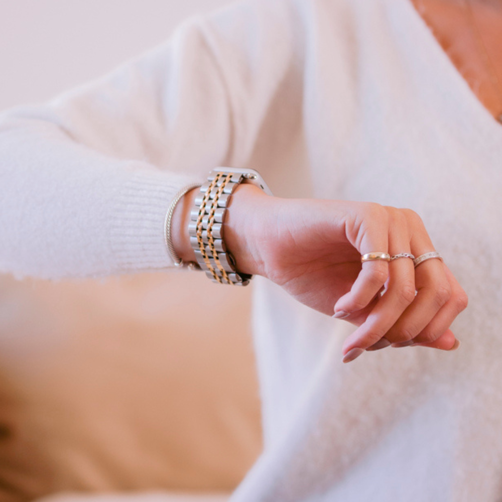 A person wearing a white long-sleeve top and multiple rings is seen adjusting a Coconut Lane Stainless Steel Apple Watch Strap - Silver & Gold on their wrist. The background is blurred, focusing on the sleek adjustable strap and the watch.