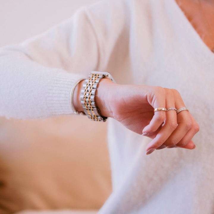 A person wearing a white long-sleeve top and multiple rings is seen adjusting a Coconut Lane Stainless Steel Apple Watch Strap - Silver & Gold on their wrist. The background is blurred, focusing on the sleek adjustable strap and the watch.