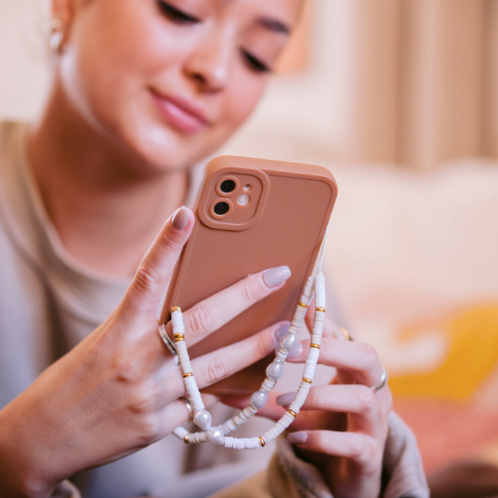 A person with light-colored nails holds a pink smartphone adorned with a beaded strap. The background is softly blurred, and the individual appears to be seated and engaged with the phone, smiling slightly, while a NAKD Phone Case - Chocolate Brown from Coconut Lane rests beside them.